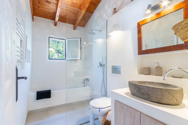 Een badkamer met een wasbak van steen, een regendouche boven het bad achter een glazen wand, en een houten balkenplafond.
