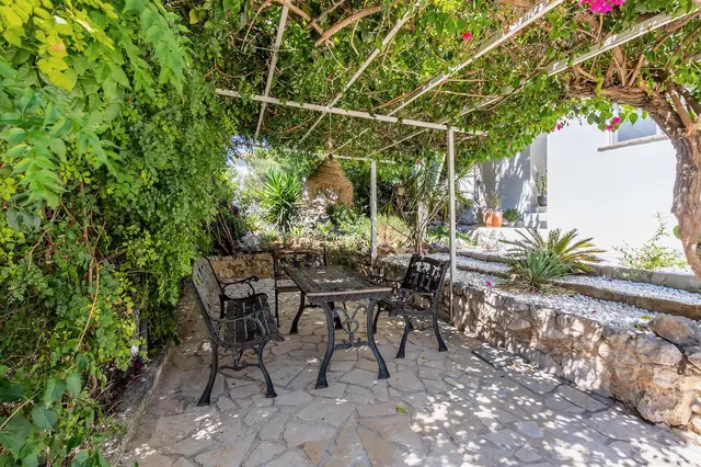 Twee banken en een stoel bij een tafel onder een dicht bladerdak van een klimplant en bougainville, op natuursteen bij een stapelmuur.