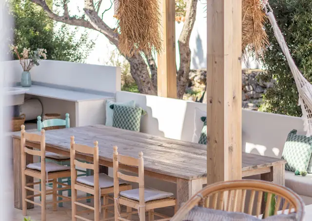 Een lange houten tafel met rieten stoelen onder de pergola, met een gemetselde bank met kussens en een wit werkblad met een spoelbak.