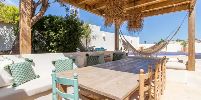De pergola van opzij, met de lange tafel, de gemetselde bank, een hangmat en het zwembad erachter.